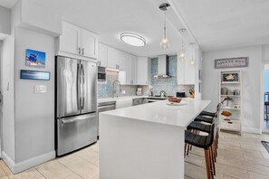 A modern kitchen with a stainless steel refrigerator, white cabinetry, a large island with bar stools, and pendant lighting.