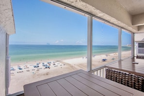 A beach view from a balcony with a table and chairs, overlooking a sandy beach with umbrellas.