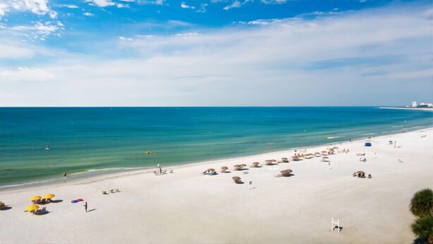 A beach with clear blue water, sandy shore, and umbrellas. - 704S Crescent Arms – Full Gulf Front View (Siesta Key)