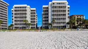 A beachfront hotel with multiple-story buildings, a sandy beach in the foreground, and palm trees. - 704S Crescent Arms – Full Gulf Front View (Siesta Key)