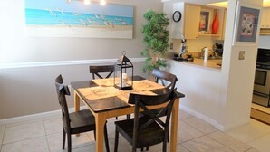 A dining area with a wooden table and chairs, a painting of a beach scene, a clock, and a kitchen area visible through an open doorway. - Ground-level Condo near Beach and Shops - Casa Siesta 2 (Siesta Key)