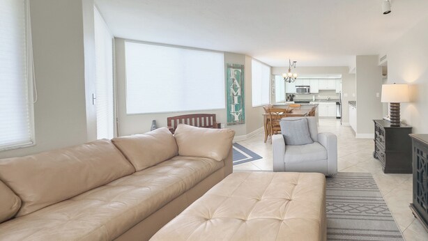 Een moderne woonkamer met een grote beige bank en een bijpassend pouf, een eethoek met een houten tafel en stoelen, en een keuken op de achtergrond.