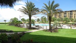 Um resort com piscina, palmeiras e um prédio grande ao fundo.
