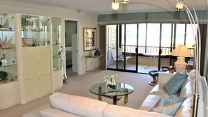 A modern living room with a glass coffee table, white sofa, and a balcony with a view. - Gulf and Bay Club- A603 (Siesta Key)