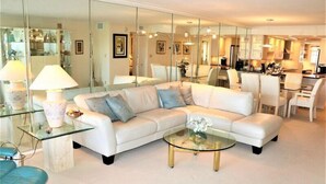 A modern living room with a white sofa, glass coffee table, and a dining area with chairs and a table. - Gulf and Bay Club- A603 (Siesta Key)