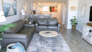A living room with a grey leather sofa, a coffee table, and a dining area with a kitchen in the background.