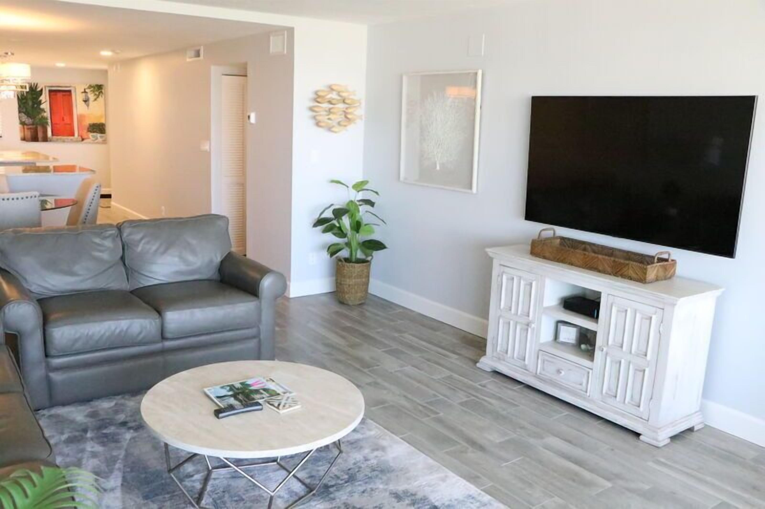 A living room with a grey leather sofa, a round coffee table, a white entertainment unit with a flat-screen TV, and a potted plant.