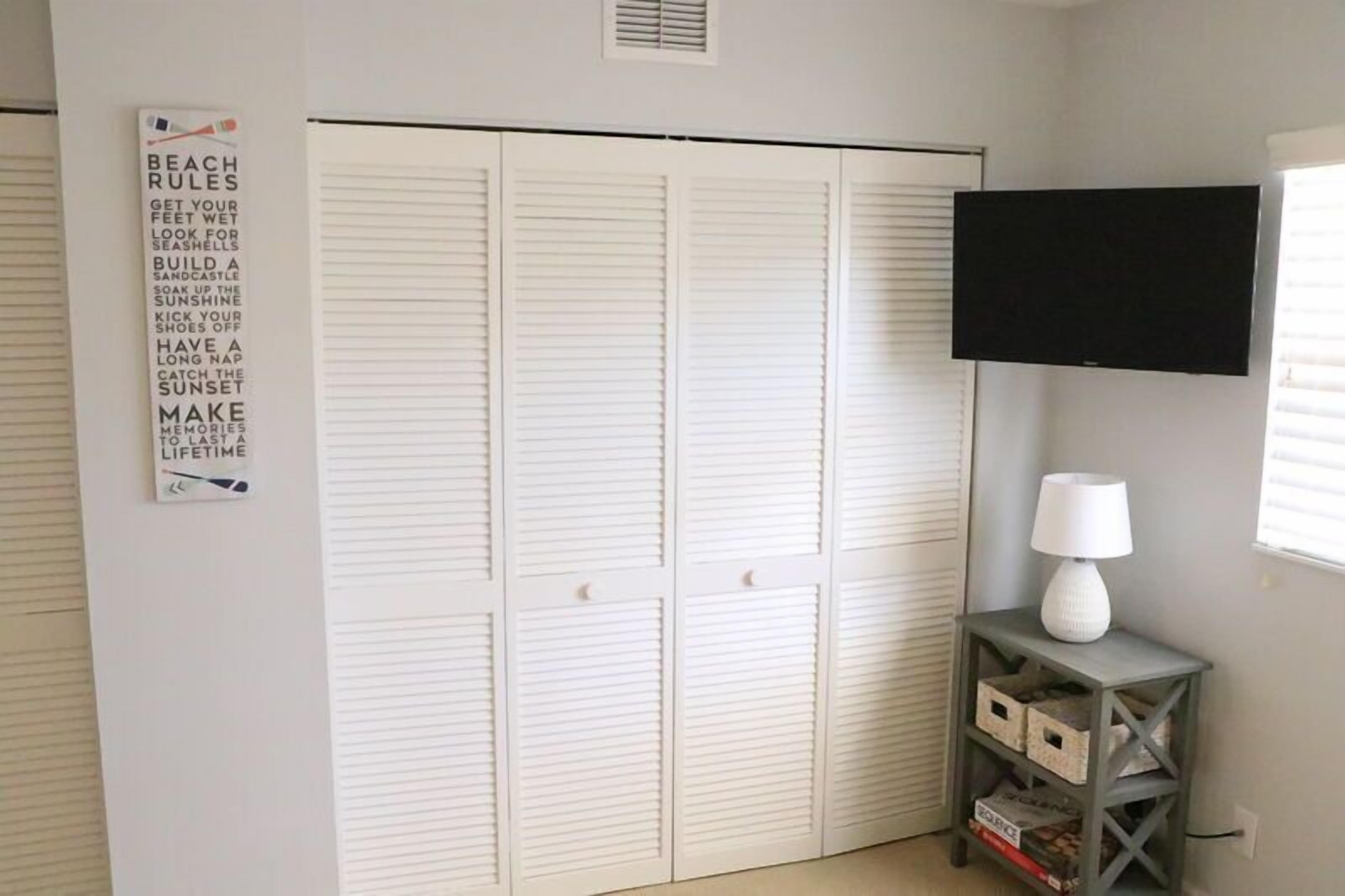 A room with a white closet, a mounted TV, a white lamp, and storage boxes.
