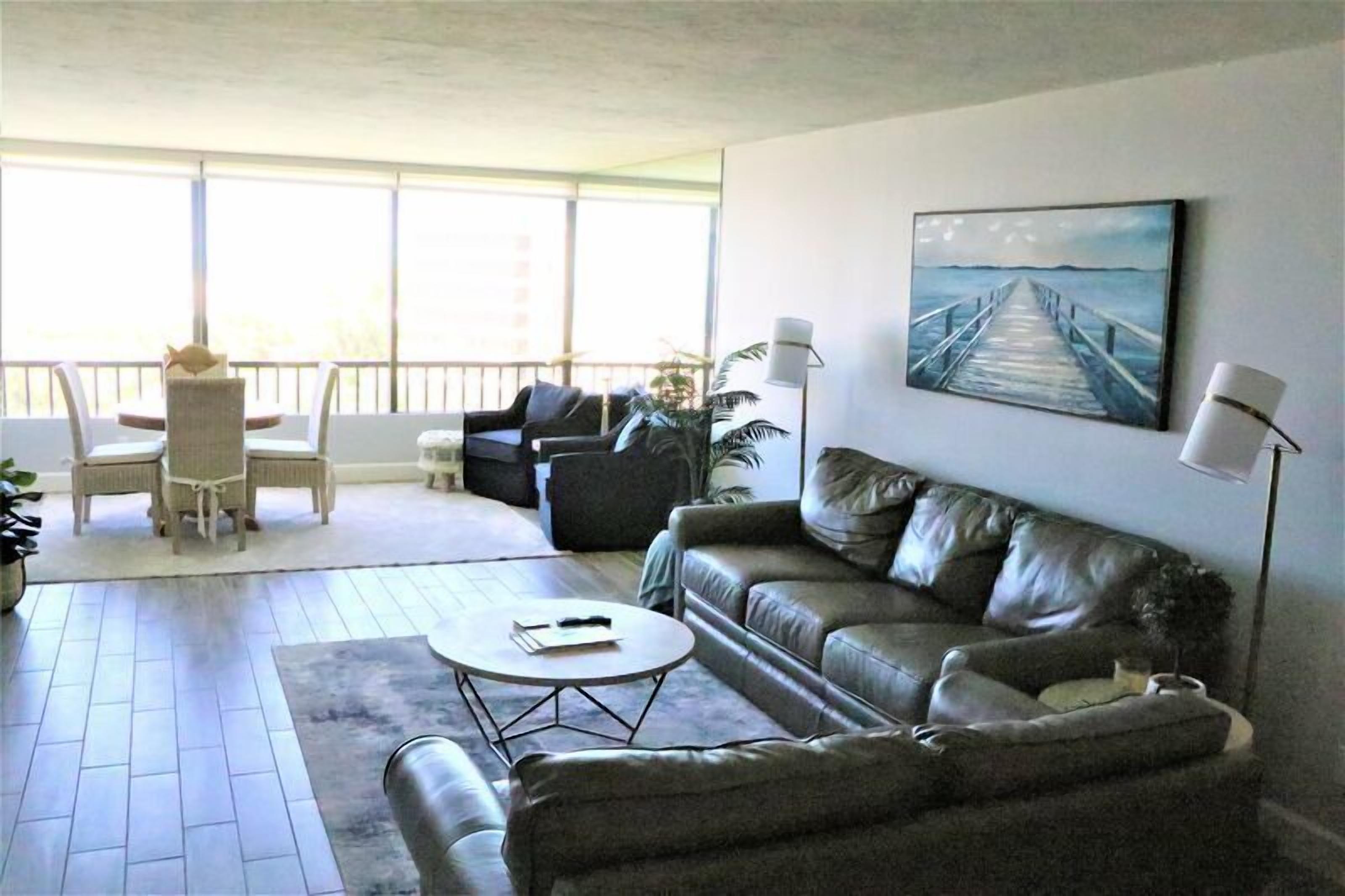 A living room with a sofa, a coffee table, and a dining area with chairs and a table. There is a large window with a view of the outside.
