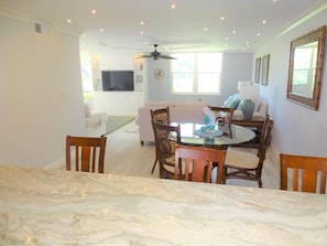 A dining area with a marble table, wooden chairs, a ceiling fan, and a living room with a sofa and TV in the background.