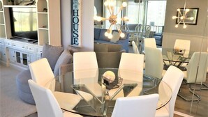 A modern living room with a glass dining table surrounded by white chairs, a built-in entertainment unit, and a cozy seating area with a sofa and armchairs. - Gulf and Bay Club- B205 (Siesta Key)