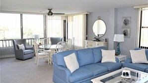 A modern living room with a blue sofa, white armchairs, a dining table, and a ceiling fan. - Gulf and Bay Club- B205 (Siesta Key)