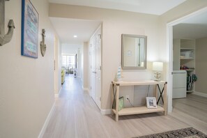 A hallway with a wooden console table, a mirror, and a framed picture. There is a door leading to another room and a small refrigerator in the corner. - Gulf and Bay Club- B301 Updated end unit! (Siesta Key)