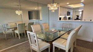 A modern dining area with a glass table and white chairs, a chandelier, and a kitchen in the background.
