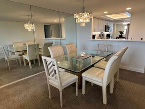 Un espace repas moderne avec une table en verre et des chaises blanches, un lustre et une cuisine en arrière-plan.