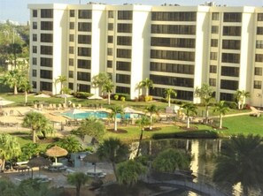 A multi-story hotel with a swimming pool, palm trees, and a small bridge.