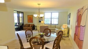 A dining area with a round table and six chairs, a sofa, a TV, and a painting on the wall. - Gulf and Bay Club- B608 Beautiful and great view! (Siesta Key)