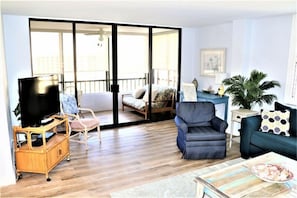 A living room with a television on a wooden stand, a blue armchair, a sofa, and a balcony with a bed.
