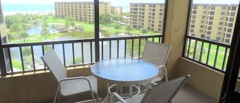 A balcony with a round table and four chairs, overlooking a landscaped area with buildings and a body of water.