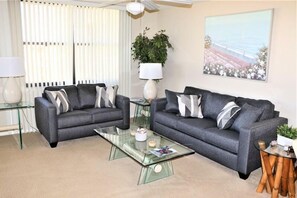 Un salon avec deux canapés gris, une table basse en verre, une lampe et un tableau au mur.