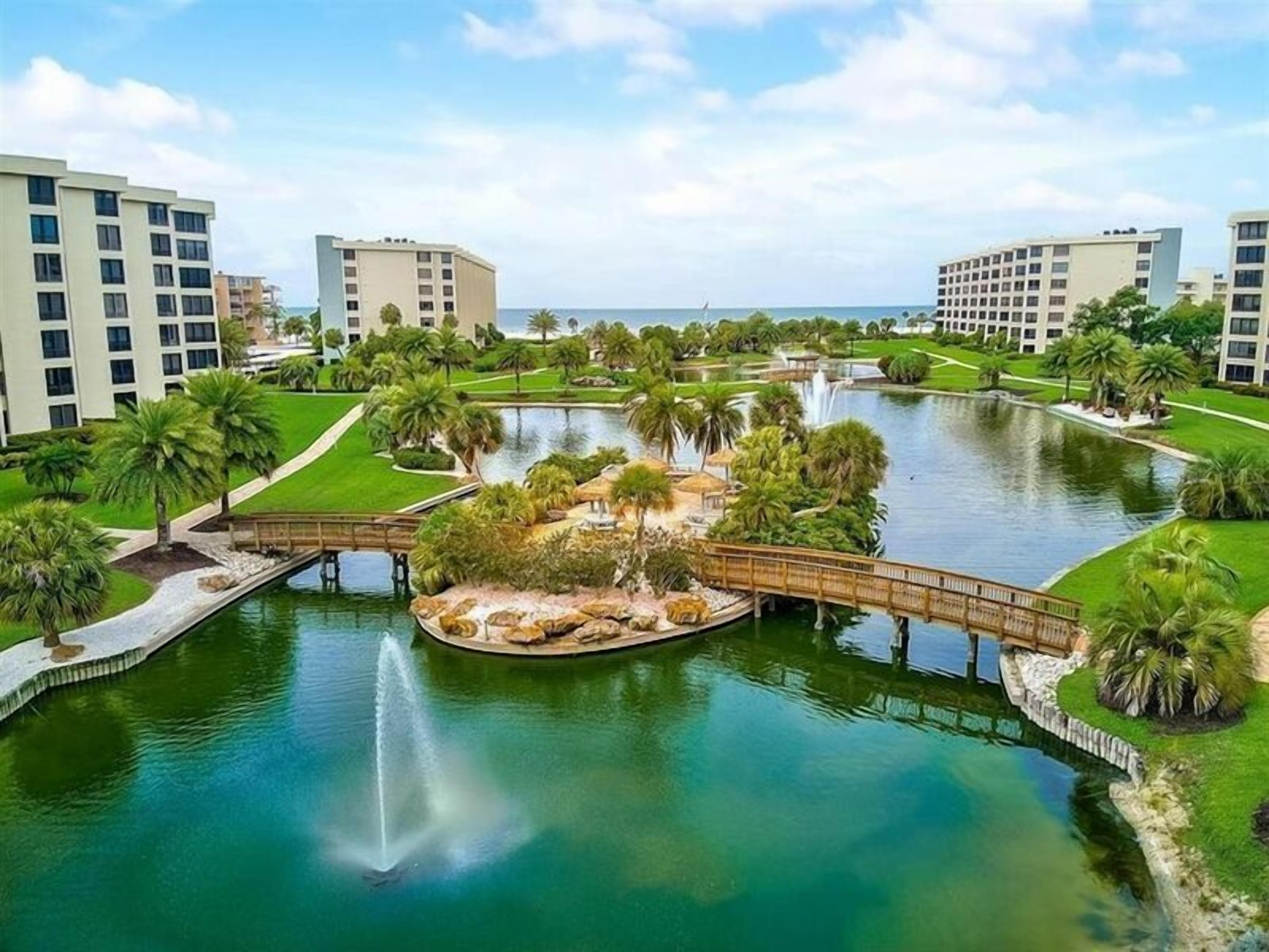 Un complejo turístico con fuente central, palmeras y un puente que conecta distintas zonas.