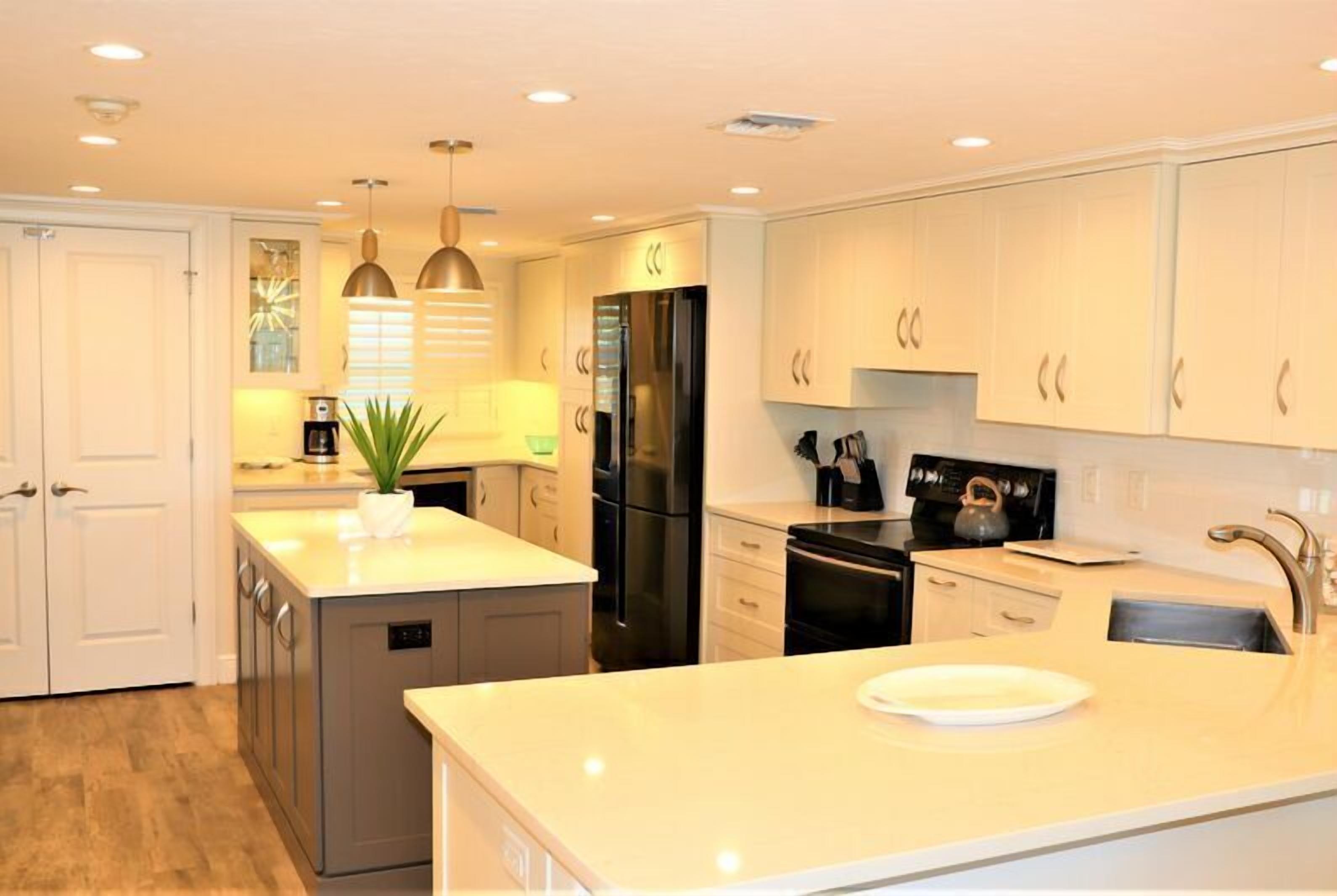 Cocina moderna con armarios blancos, una isla central, electrodomésticos de acero inoxidable, iluminación colgante y una pequeña planta en maceta.