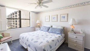 A bedroom with a bed, two bedside tables, a chair, a ceiling fan, and a window with blinds.