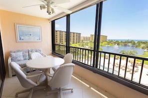 Uma varanda com mesa e cadeiras, um ventilador de teto e uma janela grande com vista para uma piscina e edifícios.