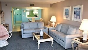 A living room with two grey sofas, a glass coffee table, and a white lamp. There is a door leading to a kitchen and a hallway.