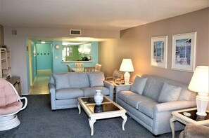 A living room with two grey sofas, a glass coffee table, and a white lamp. There is a door leading to a kitchen and a hallway.