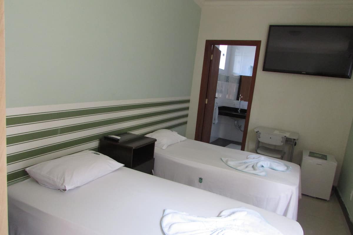 Habitación de hotel con dos camas individuales, una mesita de noche pequeña con un control remoto, un televisor de pantalla plana montado en la pared y un escritorio pequeño con una impresora.