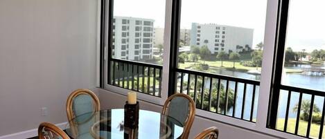 A dining area with a glass table and wicker chairs, large windows with a view of buildings and a body of water.