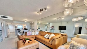 A modern living room with a sectional sofa, a coffee table, and a patterned armchair. There is a kitchen area in the background with a dining table and chairs. - Gulf and Bay Club- C702 Beautifully updated! (Siesta Key)