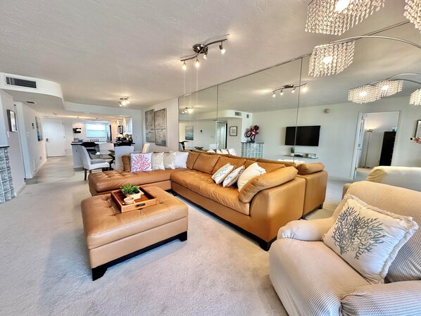 A modern living room with a sectional sofa, a coffee table, and a patterned armchair. There is a kitchen area in the background with a dining table and chairs.