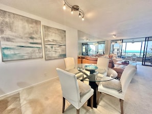 A modern living room with a glass dining table, white chairs, and a sofa. There are abstract paintings on the wall and a view of the ocean through large windows.