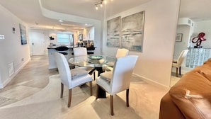 A modern dining area with a glass table and white chairs, a kitchen in the background, and abstract art on the walls.