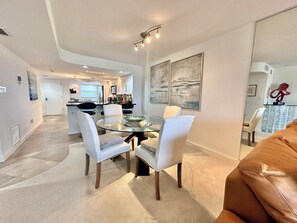 A modern dining area with a glass table and white chairs, a kitchen in the background, and abstract art on the walls.