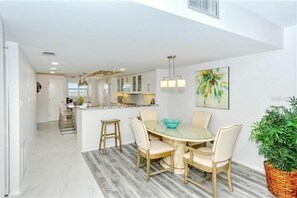 Una cocina moderna con armarios blancos, una isla central y un comedor con una mesa de vidrio y sillas.
