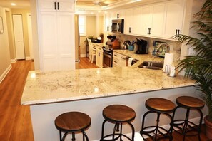 A kitchen with a large island, white cabinets, and a microwave. There are three stools at the island. - Gulf and Bay Club- D209 (Siesta Key)