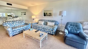 A living room with a blue patterned sofa set, a matching armchair, a coffee table, and a dining area in the background. - Lanai and Beach Acess - Gulf and Bay Club E104 (Siesta Key)
