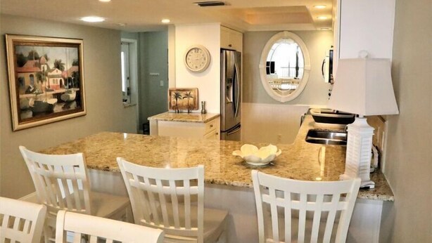 A kitchen with a granite countertop, white chairs, a refrigerator, and a clock on the wall. - Gulf and Bay Club- E108- Ground Floor- Walk Out! (Siesta Key)