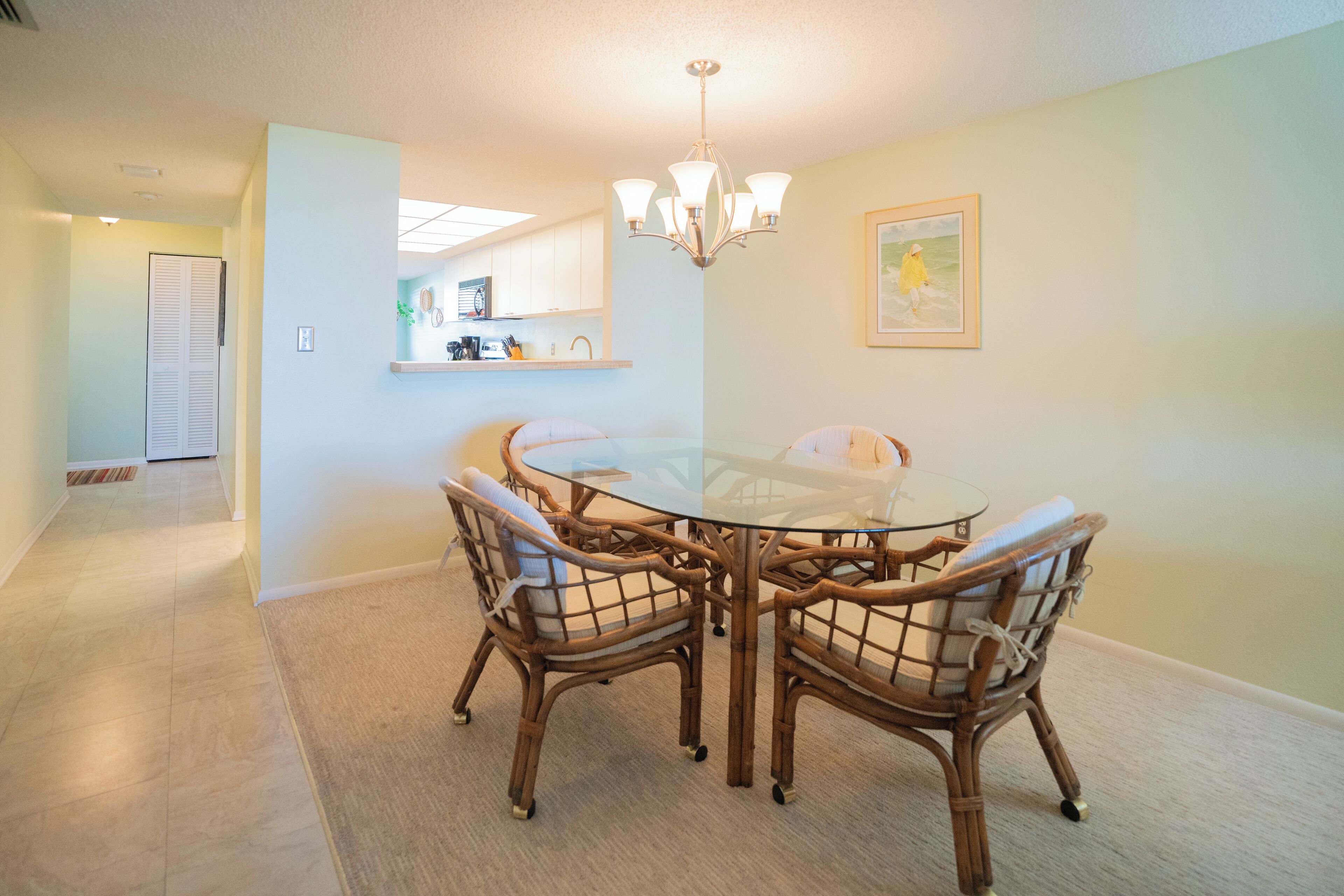 A dining area with a round glass table surrounded by wicker chairs, a chandelier, and a kitchen in the background.