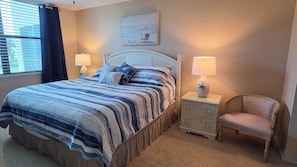 A bedroom with a bed, two bedside tables with lamps, a chair, a window with curtains, and a painting on the wall.