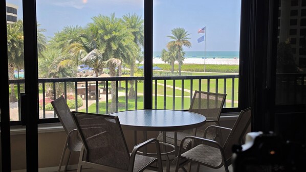 A balcony with a table and chairs, overlooking a beach with palm trees and a flag.