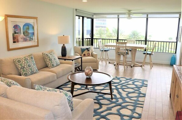 A living room with a sofa, coffee table, and chairs. There is a balcony with a table and chairs. - Gulf and Bay Club- E308 Beautiful unit with awesome views! (Siesta Key)