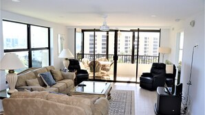 Ein geräumiges Wohnzimmer mit einer Sofagarnitur, einem Couchtisch und einem Fernseher. Ein Balkon bietet Blick auf die umliegenden Gebäude.