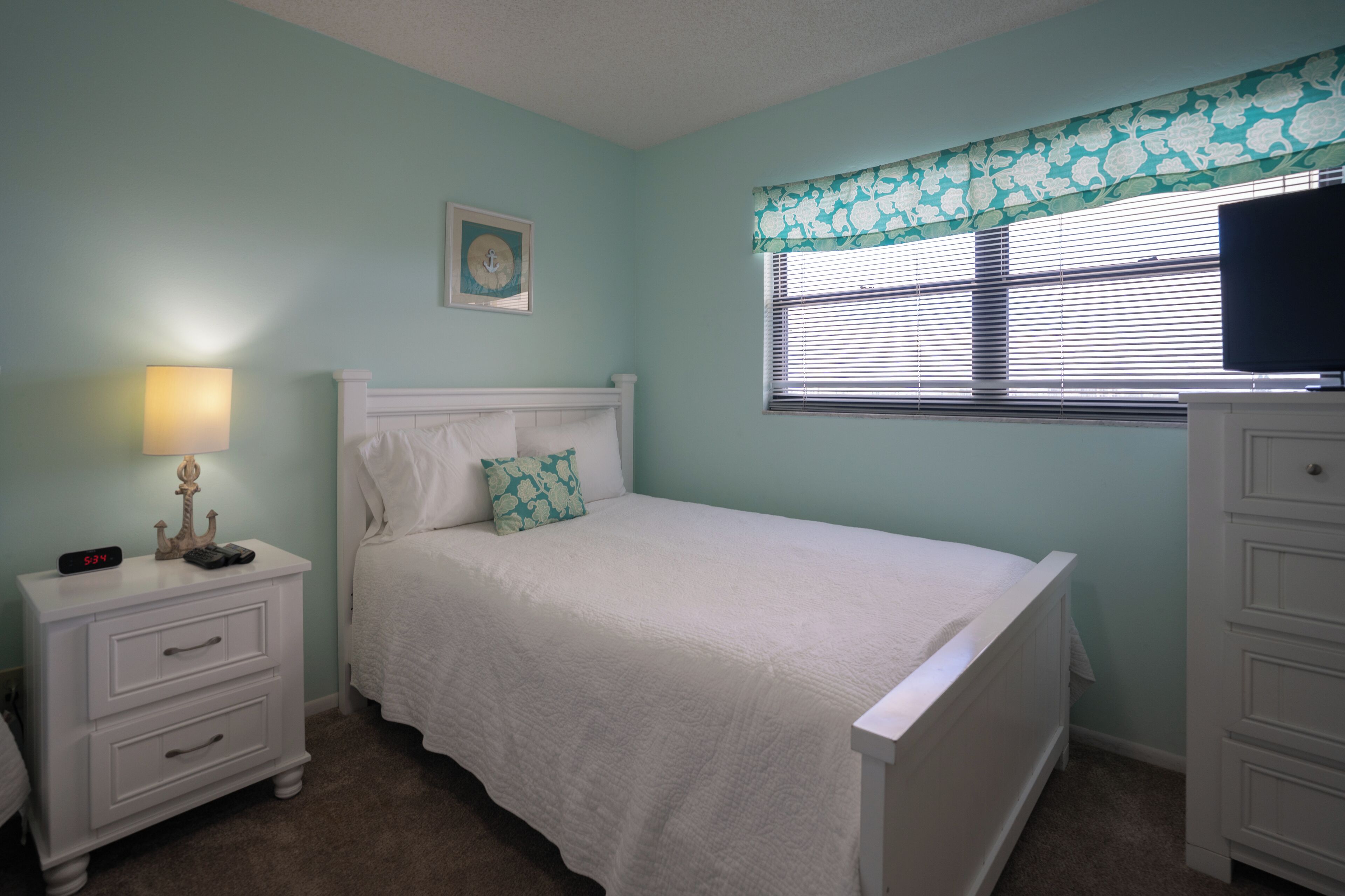 A bedroom with a bed, a nightstand, a dresser, a window with blinds, and a wall clock.