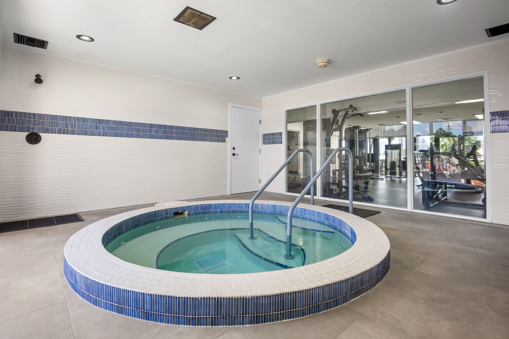 A circular hot tub with a tiled edge, surrounded by a tiled floor and walls, with a door and a view of a gym area.