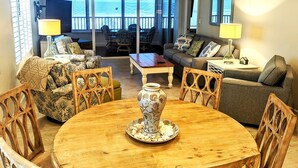 A spacious living room with a round wooden table, comfortable seating, and a large window offering a view of the outdoors.