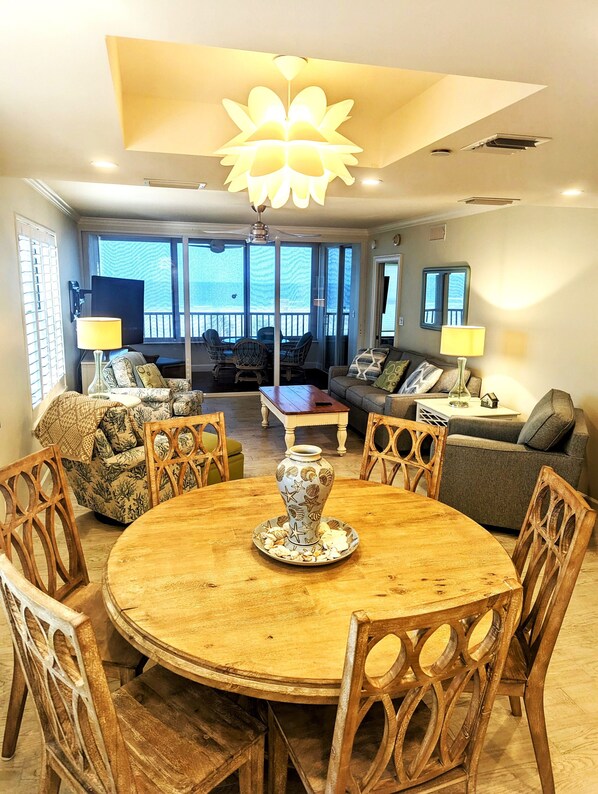 A spacious living room with a round wooden table, comfortable seating, and a large window offering a view of the outdoors.
